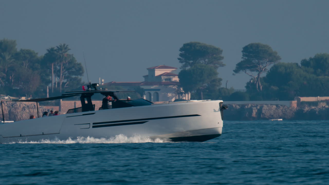 Cannes, France - October 10, 2025: A white sailboat glides near the rocky coastline of the French Riviera, with lush pine trees and elegant seaside villas in the background