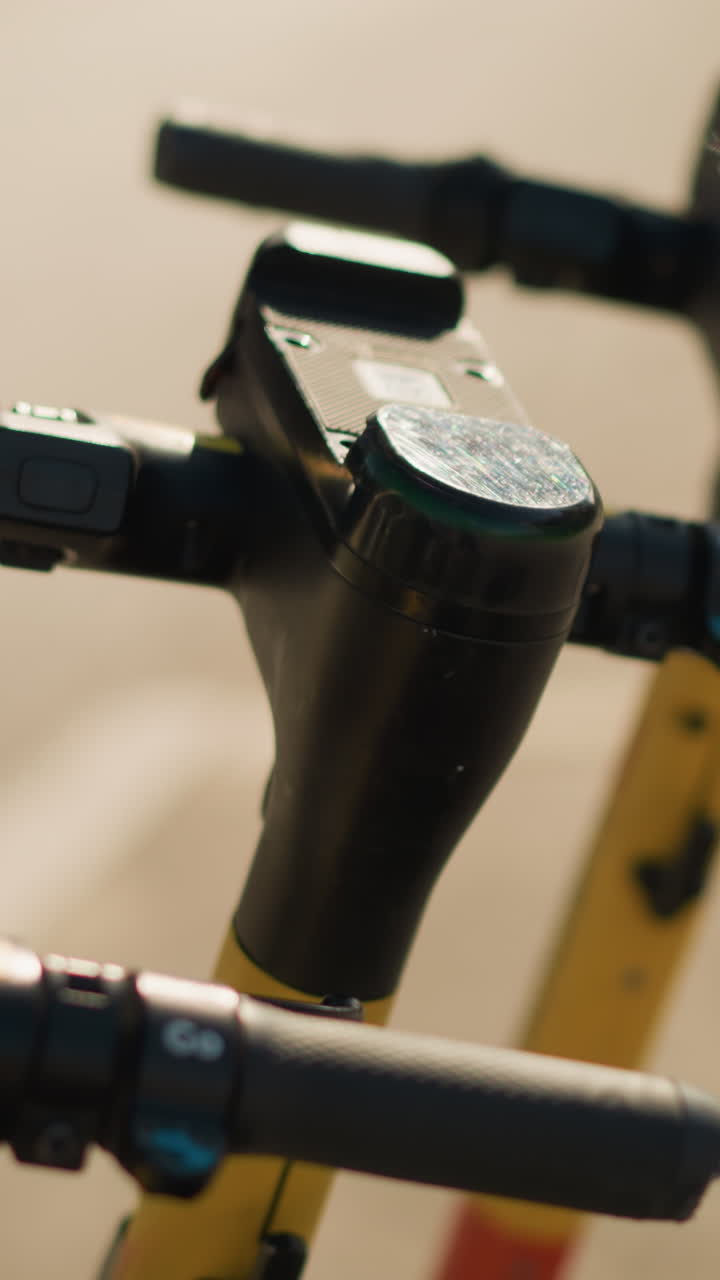 햇빛이 비치는 노란색 스템과 질감 있는 그립이 있는 전동 스쿠터 핸들바 콘솔의 클로즈업 사진입니다. 콘솔 상단에는 낡은 스티커가 붙어 있는 무인 대여 스쿠터가 있으며, 흐릿한 도시 거리 배경과 따뜻한 아침 햇살이 느껴집니다.