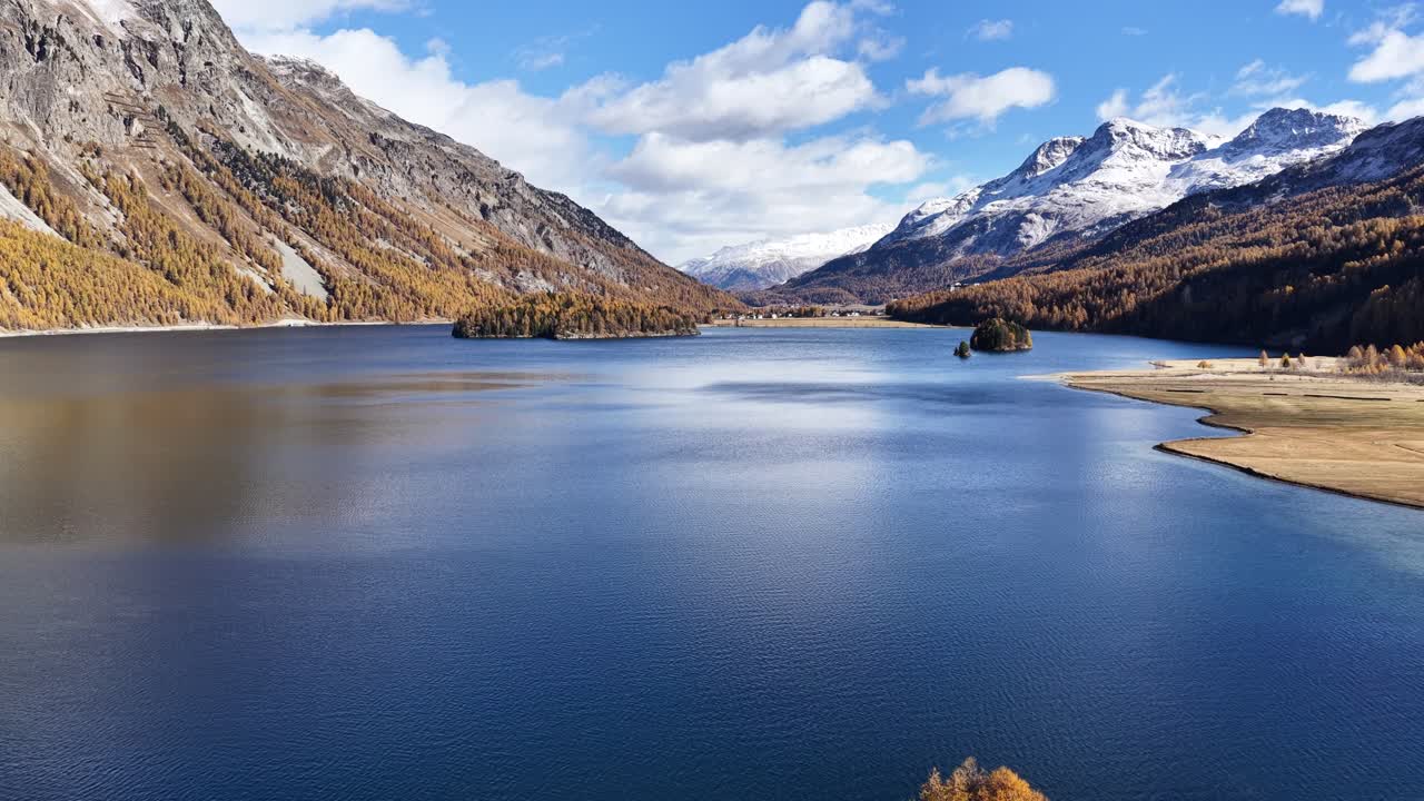 Aerial view of the clear blue water of a lake Silvaplanain Kanton Graubünden, Schweiz. The golden-yellow larches frame a view of snow-dusted peaks reflected perfectly in the tranquil water