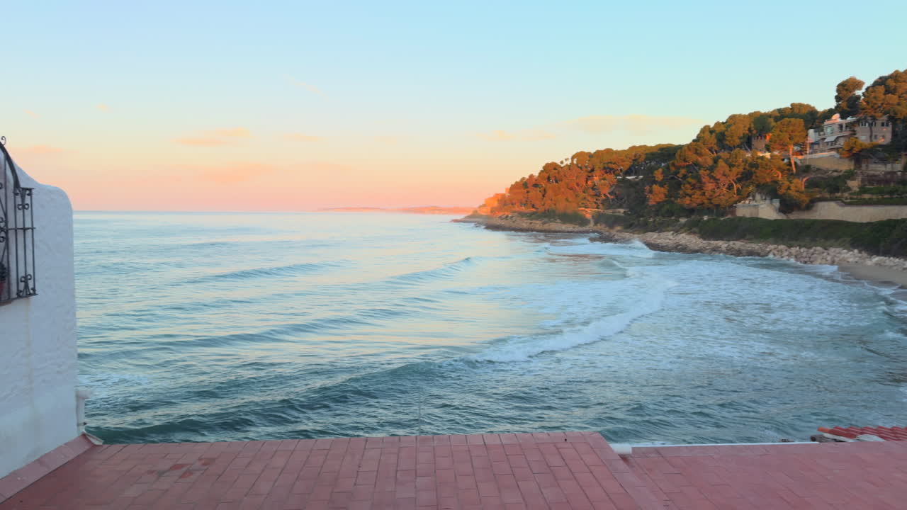 Scenic seaside view at sunset from a whitewashed building in Roc de Sant Gaietà, Spain. Gentle waves and orange light over pine-covered cliffs create a peaceful Mediterranean setting