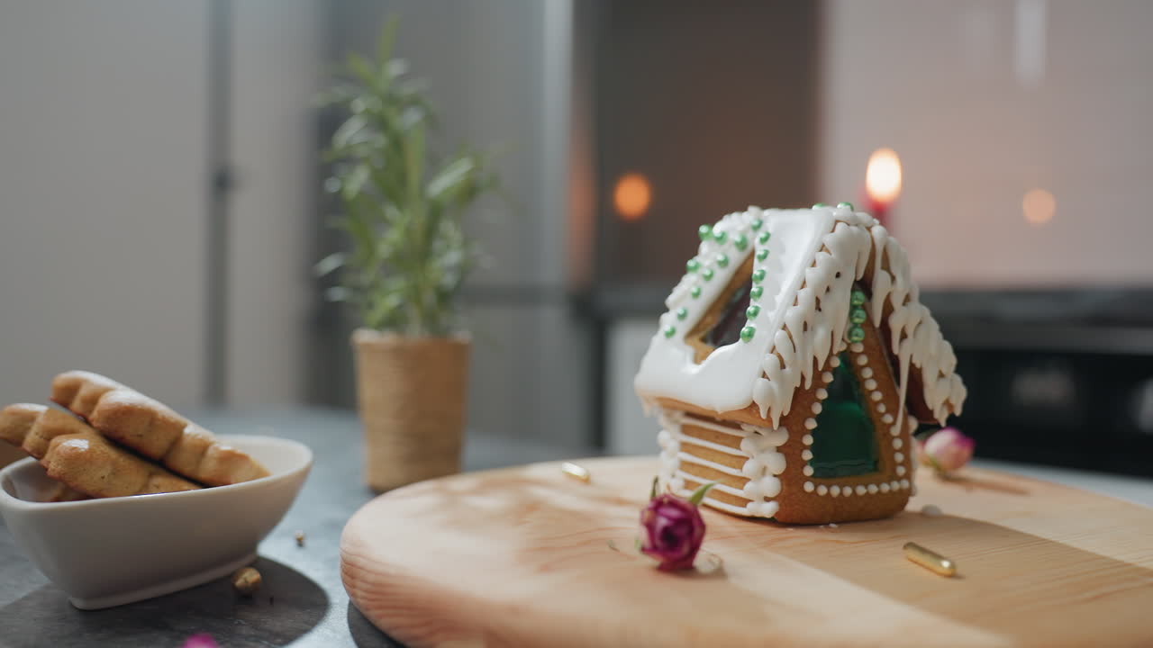 casa de pan de jengibre bellamente decorada con hielo blanco y cuentas verdes en bandeja de madera, acompañada de galletas en cuenco y planta en olla, luz de vela cálida en el fondo