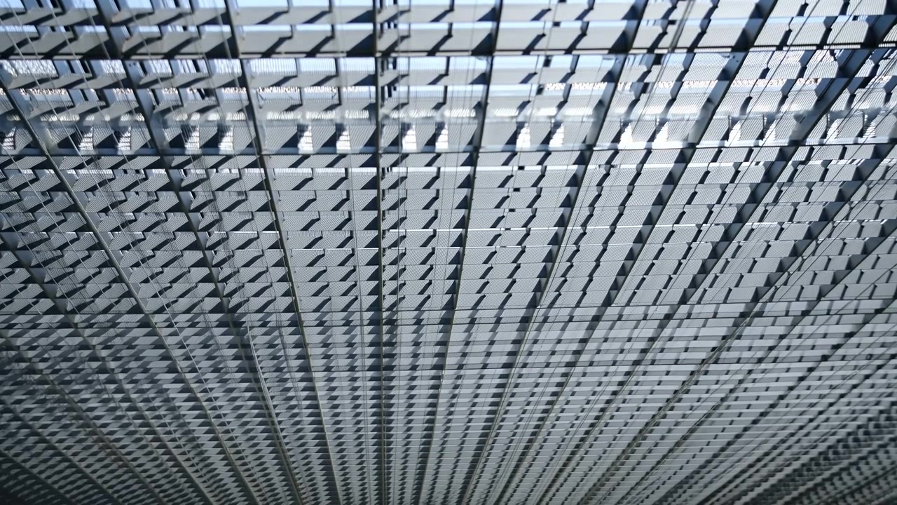 Vista architettonica dal basso che mostra persiane metalliche sospese, lamelle rettangolari e griglia di supporto che formano linee ripetute, superfici grigio freddo, ombre sottili e una spaziosa profondità geometrica.