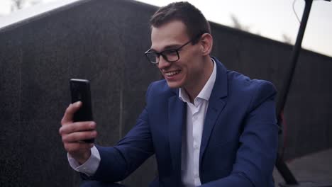 Un-Hombre-De-Negocios-Emocionado-Y-Sonriente-Sentado-En-Las-Escaleras-Y-Haciendo-Video-Chat-En-Un-Teléfono-Inteligente-Afuera.-Hombre-Con-Elegante-Traje-Azul-Y