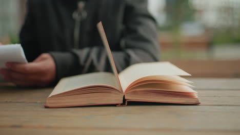 Close up of open book with pages swaying in wind on wooden table while partial view of person writes in notebook, capturing calm outdoor study atmosphere with natural movement