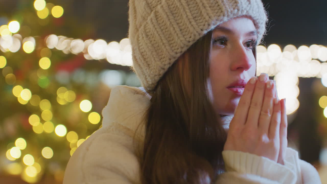 Close-up of woman bringing elegant hands to mouth, exhaling warm breath in cold winter night, visible breath vapor, blurred background features passerby and festive golden lights