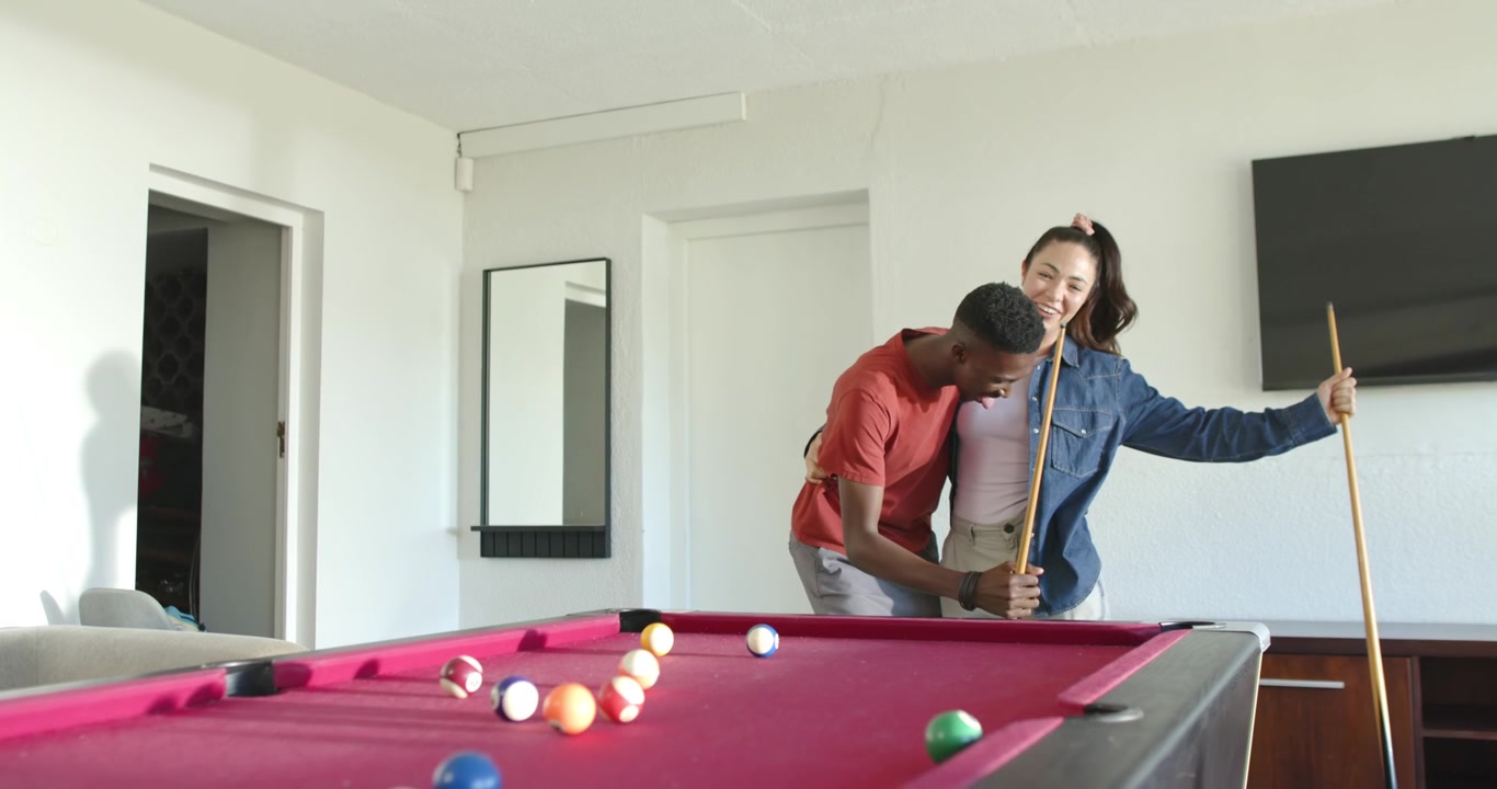 Diverse couple chatting, man stepping closer teaching her cue grip at pool table with racked balls