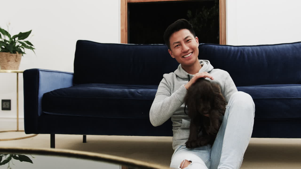 Smiling Asian man sitting on floor at home, petting adorable dog