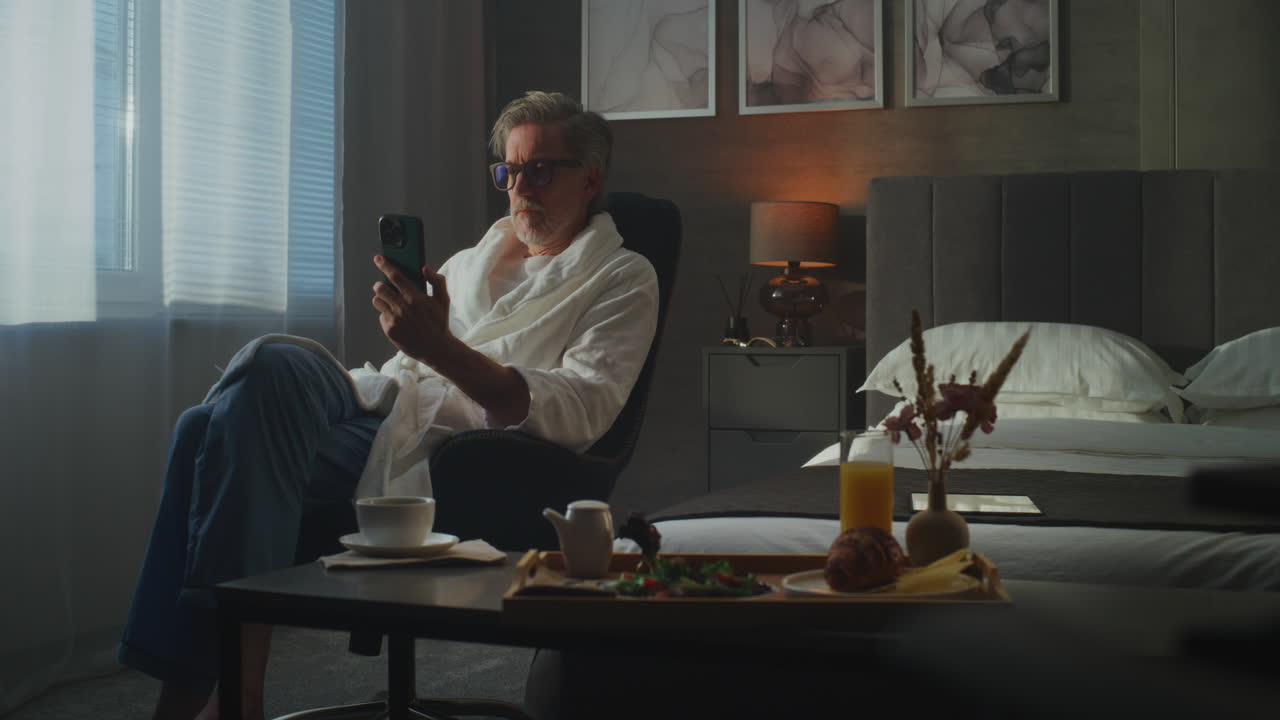 Stylish Senior Man in Soft White Bathrobe Uses Smartphone to Shop Online and Manage Payments (Stock Footage)