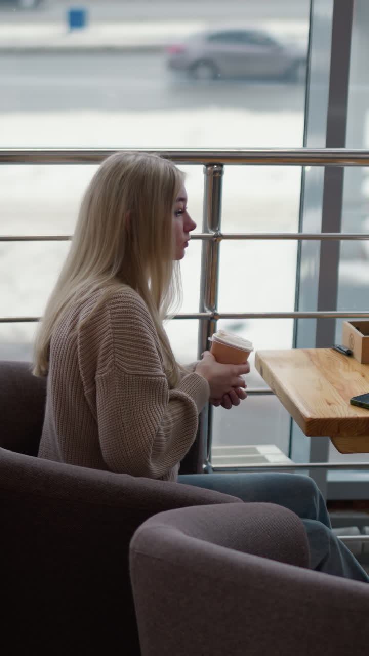 joven con una taza de café, bebiendo de ella en un café del centro comercial con una vista borrosa de la ciudad a través de la ventana, el teléfono colocado en la mesa con una atmósfera serena