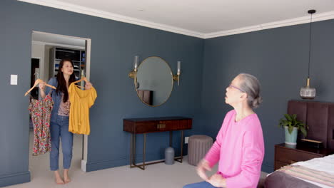 asian grandmother and granddaughter holding clothes on hangers in bedroom