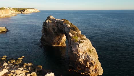 Breathtaking-aerial-orbit-of-scenic-rock-formation-Es-Pontàs-facing-calm-turquoise-ocean-waters-in-Spain-during-dusk