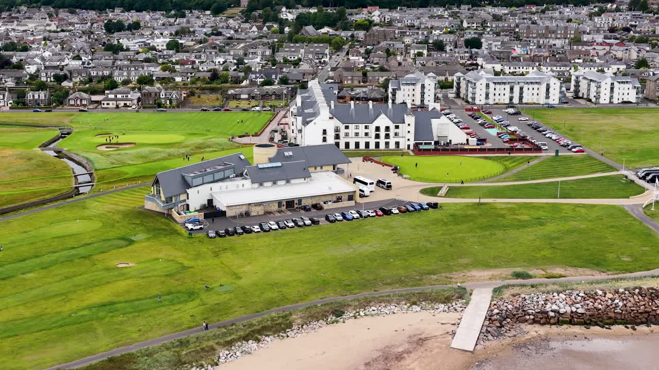 Drone pans above golf clubhouse, putting green, and coastline with town buildings in daylight