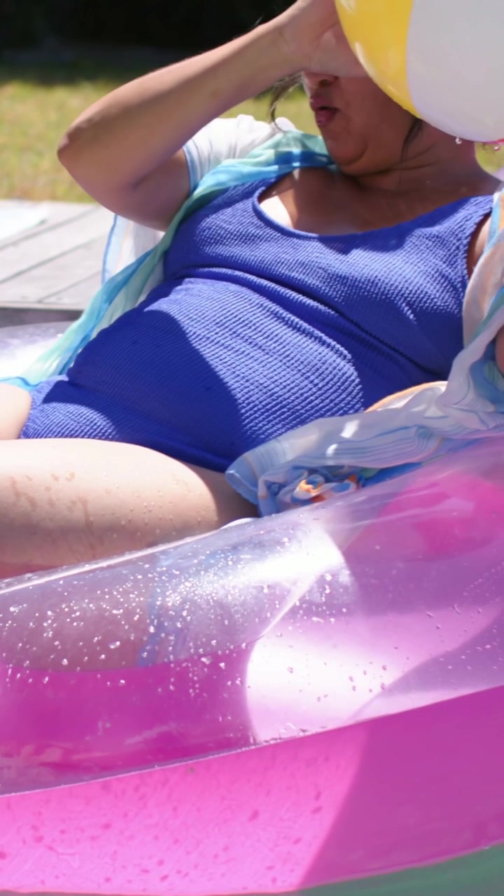 Vertical video: Lifting ball to face, woman in blue swimsuit on pink float by pool, protecting face