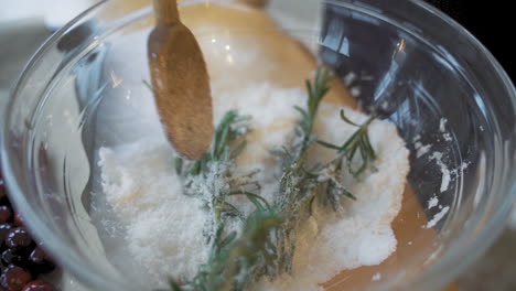 slow motion wooden spoon stirring and mixing fresh green rosemary and white sugar in large clear glass bowl on wooden table