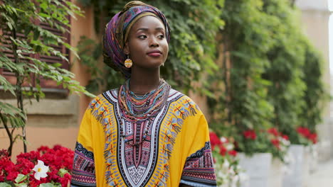 Retrato-De-La-Hermosa-Y-Feliz-Mujer-Africana-Con-El-Traje-Tradicional-Parada-En-El-Acogedor-Patio-Con-Flores-Y-Sonriendo-A-La-Cámara