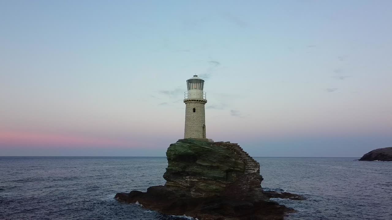 Closeup of Tourlitis Lighthouse with spiral staircase on a rocky islet from the port of Chora at sunset, Greek island of Andros, Aegean Sea, Drone shot
