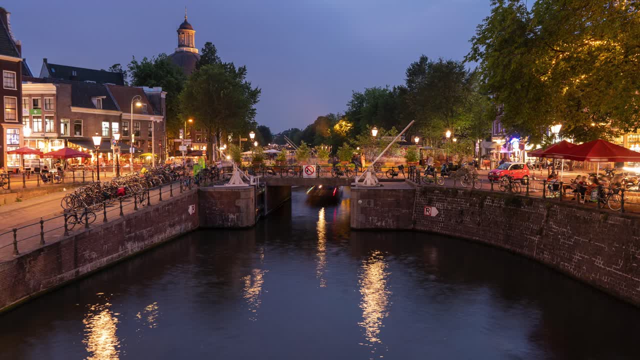 Timelapse panorámico de una intersección concurrida con peatones, ciclistas, tráfico y barcos en un canal en Ámsterdam de noche.
