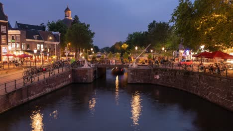 A-wide-timelapse-of-a-busy-intersection-with-pedestrians,-cyclists,-traffic-and-boats-on-a-canal-in-nighttime-Amsterdam