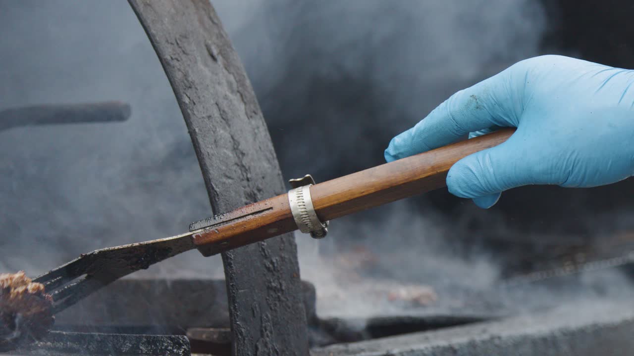 A person wearing blue gloves uses a spatula to flip burgers on a smoky barbecue grill outdoors. Natural lighting, close-up perspective, visible smoke, and steady camera movement