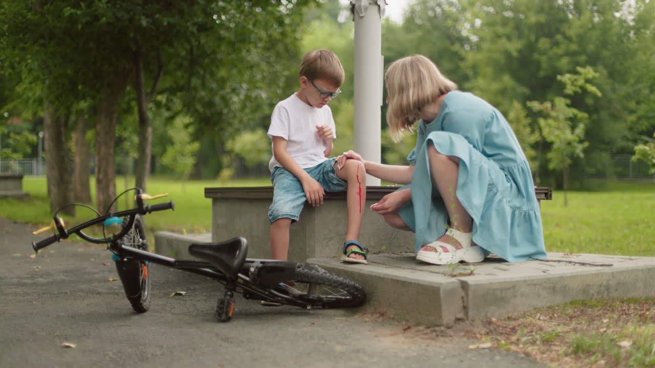 una madre preocupada se arrodilla al lado de su hijo herido, limpiando una herida sangrienta en la rodilla con un pañuelo, mientras el niño persigue moscas de la herida