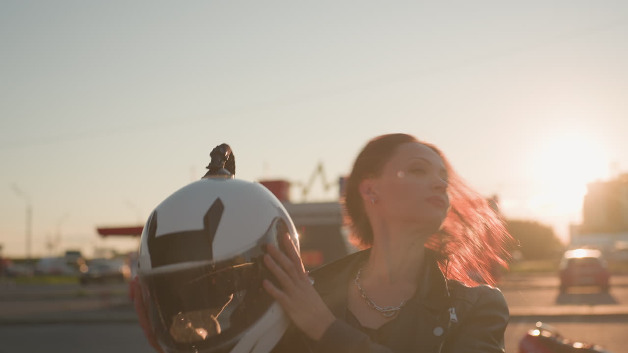 Young lady removes helmet with focused look wearing leather jacket in front of urban buildings during sunset showing determination strength lifestyle and freedom