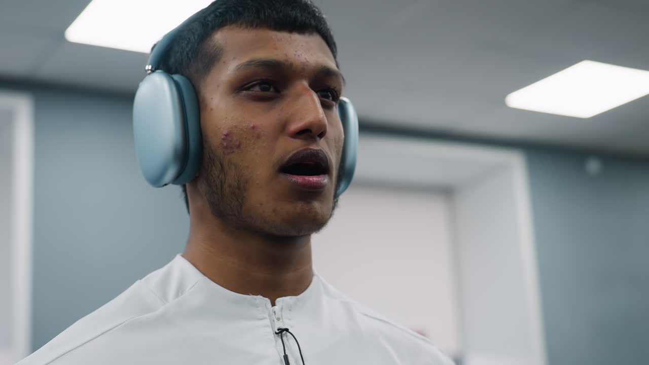 close up of focused man wearing over ear headset and white zip-up top puffing cheeks while exerting visible pressure during barbell lift in well lit gym with blurry door and ceiling lights behind