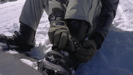 snowboarder strapping on a snowboard binding while sitting in the snow on the mountainside