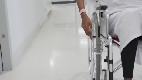 midsection of african american female patient in wheelchair in hospital corridor, slow motion