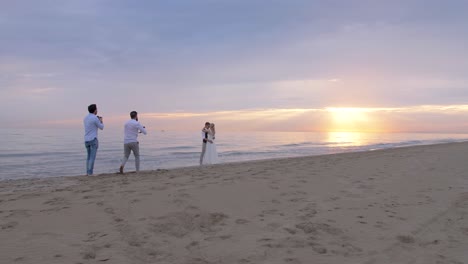 wide shot of the beach with the sun setting over the sea, of two photographers shooting a newlywed couple during their wedding