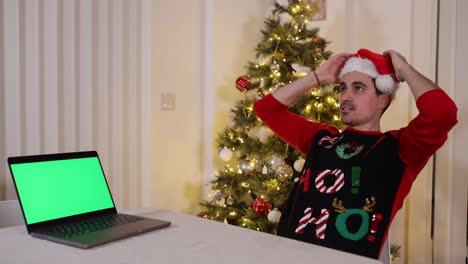 tipo con sombrero de santa y suéter recostado en una silla con una computadora portátil de pantalla verde sintiéndose contento feliz y pensando en lo que ha visto