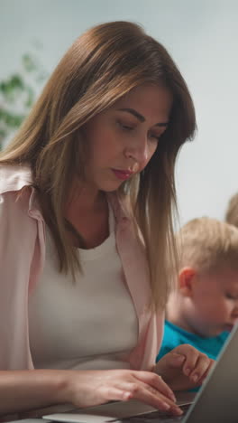 young woman in bright room works at home remotely at laptop, children sit next to their mother, learn lessons, close-up