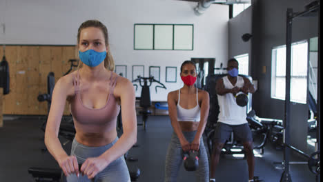 two fit caucasian women and fit african american man wearing face masks exercising using kettlebell