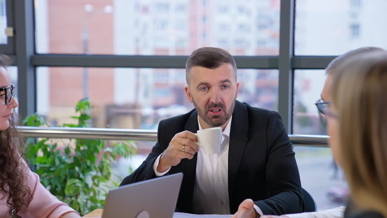 Businessman in the head of the table addresses his colleagues and drinks coffee. Young people listen to him carefully. Multi-storied buildings at backdrop in blur.