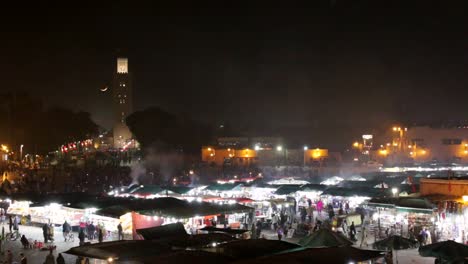 Luftaufnahme-Des-Nachtmarktes-Von-Marrakesch,-Marokko,-Mit-Touristen-Und-Einheimischen,-Die-Durch-Die-Souks-Und-Verkaufsstände-Spazieren,-Um-Essen-Und-Souvenirs-Zu-Kaufen,-Während-Die-Mondsichel-über-Der-Moschee-Aufgeht