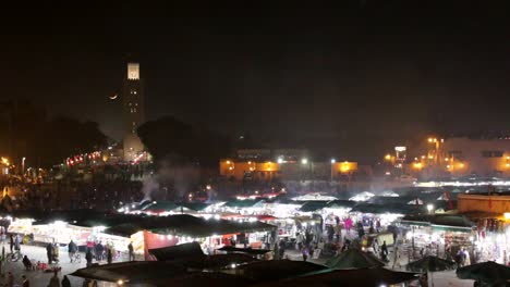 vista aérea del mercado nocturno de marrakech marruecos con turistas y lugareños caminando por los zocos y puestos de vendedores de comida y recuerdos con la luna creciente sobre la mezquita