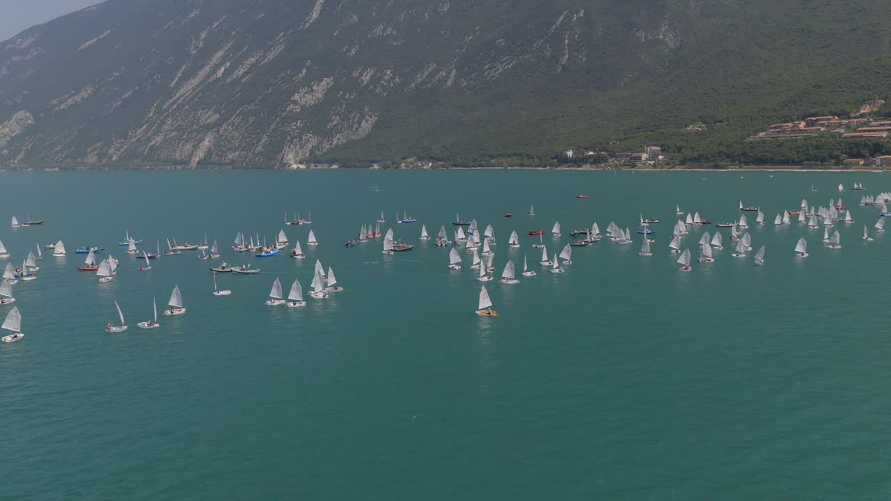 Ripresa cinematografica con drone in orbita che cattura una dinamica regata velica sul Lago di Garda, Italia. Molteplici barche a vela gareggiano sull'acqua cristallina e blu mentre il drone sorvola fluidamente la flotta, creando una scena drammatica.