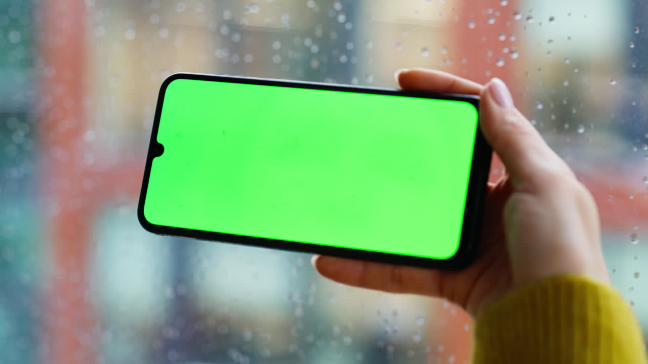 Woman finger swiping chromakey smartphone standing by rainy window closeup