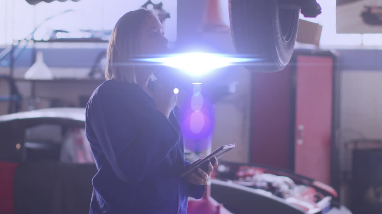 female mechanic examining lifted vehicle in auto shop, with animated tablet data and lens flare