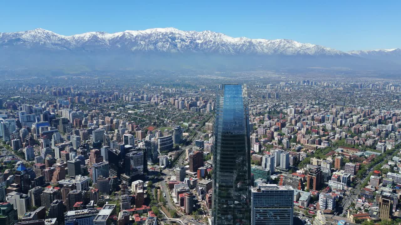 Drone aerial backward pullback revealing Costanera skyscraper and sprawling cityscape with snowcapped Andes on the horizon under clear daytime light. Santiago, Chile