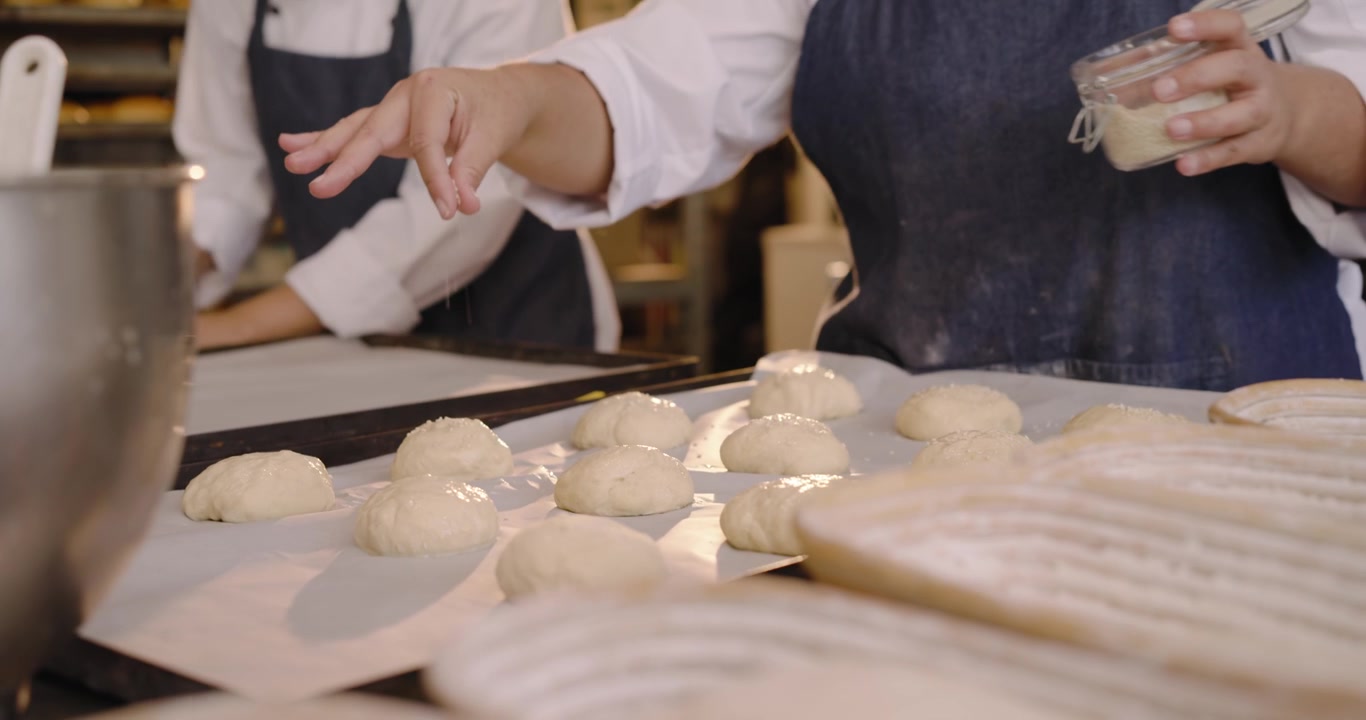 Bakers are pulling pieces from dough and shaping rounds on floured bench, glazing for proofing