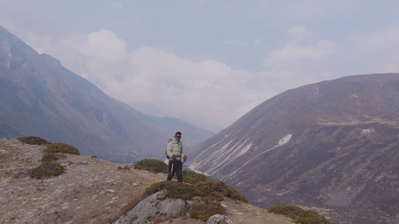 Tourist walks with trekking sticks at Dingboche, Everest Base Camp route. Cloudy hills and serene landscape create adventurous trekking visuals in Nepal