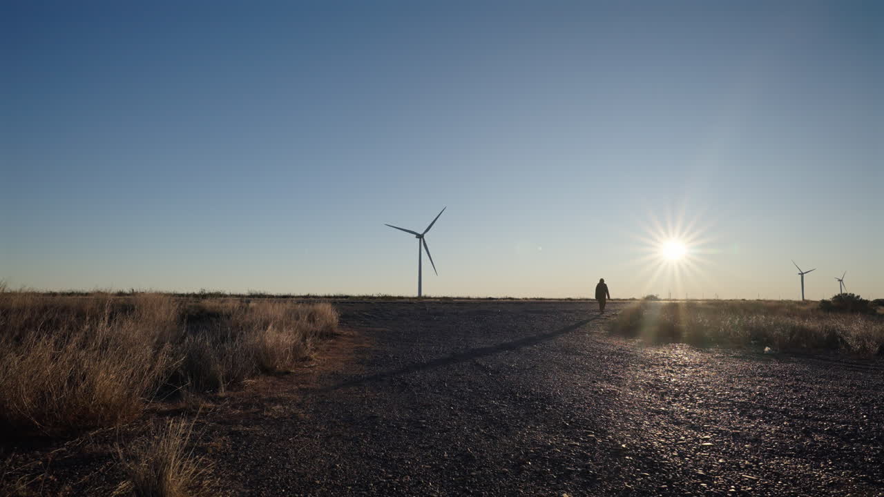 Woman walking to wind turbines while those standing on dry field open space at sunset