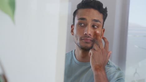 Hombre-Birracial-Mirando-En-El-Espejo-Y-Aplicando-Crema-Facial-En-El-Baño,-Cámara-Lenta