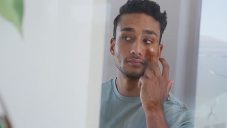 hombre biracial mirándose en el espejo y aplicando crema facial en el baño, cámara lenta