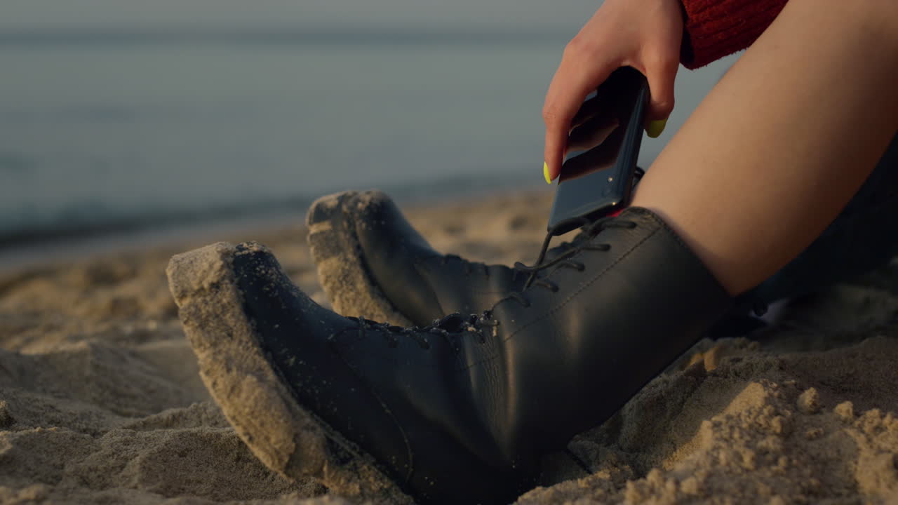Woman legs in boots sitting on sandy beach. Stylish girl holding smartphone