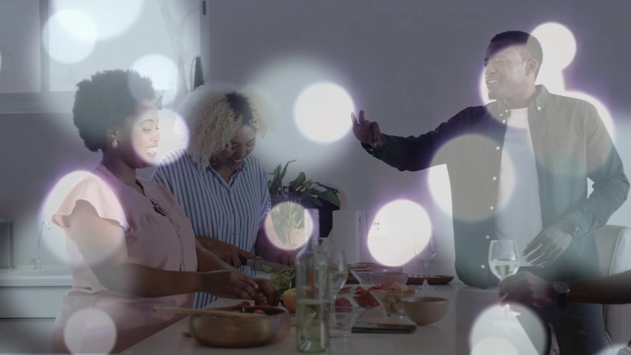 Mulheres preparando uma refeição na cozinha enquanto um homem inicia uma conversa, gesticulando, segurando vinho, com efeito bokeh.
