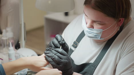 nail technician in black gloves carefully using nail tool to clean off filing residue from customer's fingernails during detailed manicure session in professional beauty salon