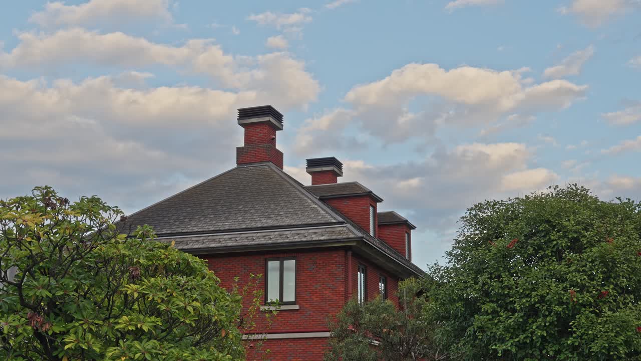 The top of a stately red brick building featuring multiple chimneys, seen against a blue sky with fluffy clouds
