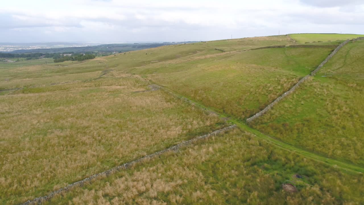 Slow moving reversing drone footage over the hills of West Yorkshire countryside, farmland and moorland flying parallel to a farmer's track and over a dry stone wall with towns in the far distance