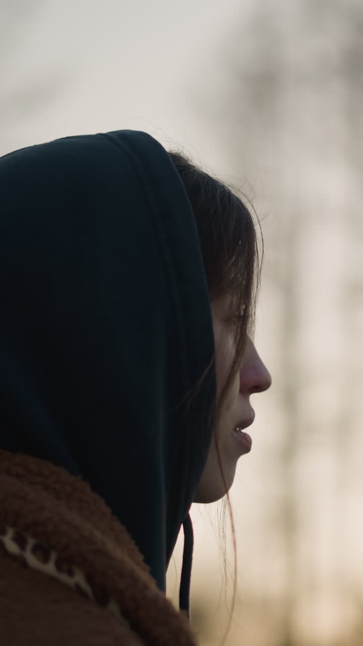 A melancholic girl walks through a park at sunset, wearing a brown coat and a hoodie with the hood up, partially concealing her face. blurred background of trees and fading sunlight